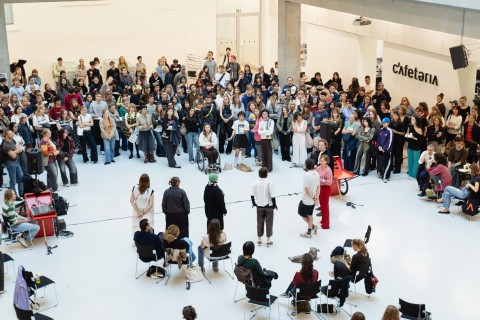 Foto der Begrüßung der Erstsemestrigen, fünf Redner*innen stehen vor einer großen Gruppe Studierender im Atrium
                           der Universität für angewandte Kunst Wien