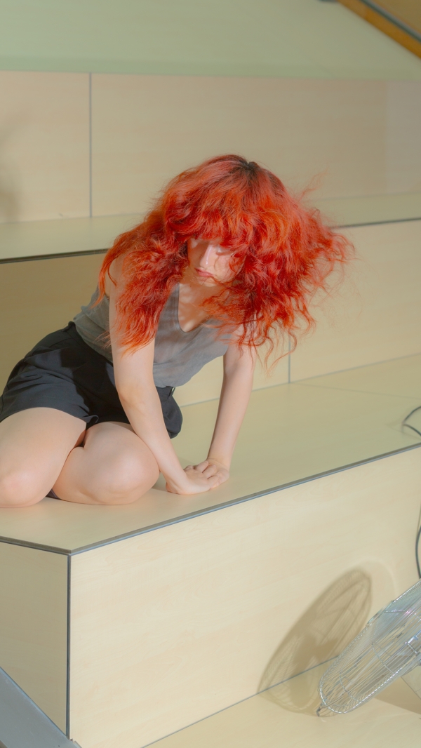 Design: 101; Foto: Luca Celine Müller Eine Person mit langen roten
                                          Haaren kniet auf einer hölzernen Plattform.