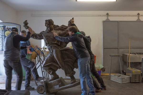 A monument of a male figure is being transported by a group of men with a hand truck.
