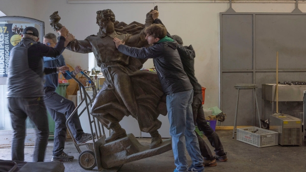 A monument of a male figure is being transported by a group of men with a hand truck.