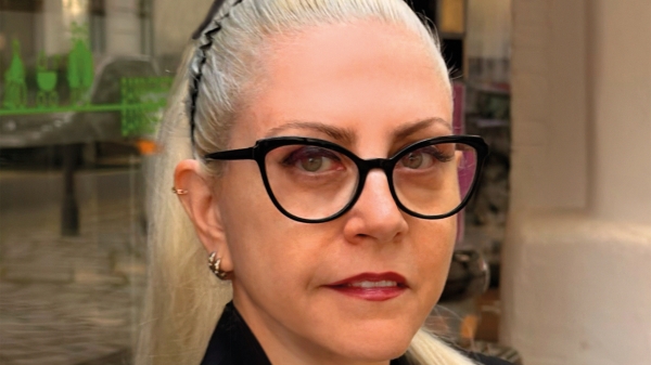 Fotoporträt
                                          ovn Basak Senova mit blondem, zurückgebundenem Haar und schwarzer Brille. Sie trägt ein schwarzes Sakko und eine Halskette
                                          und blickt direkt in die Kamera. Im Hintergrund ist ein Ausstellungsraum mit Glasflächen und Kunstwerken zu sehen.